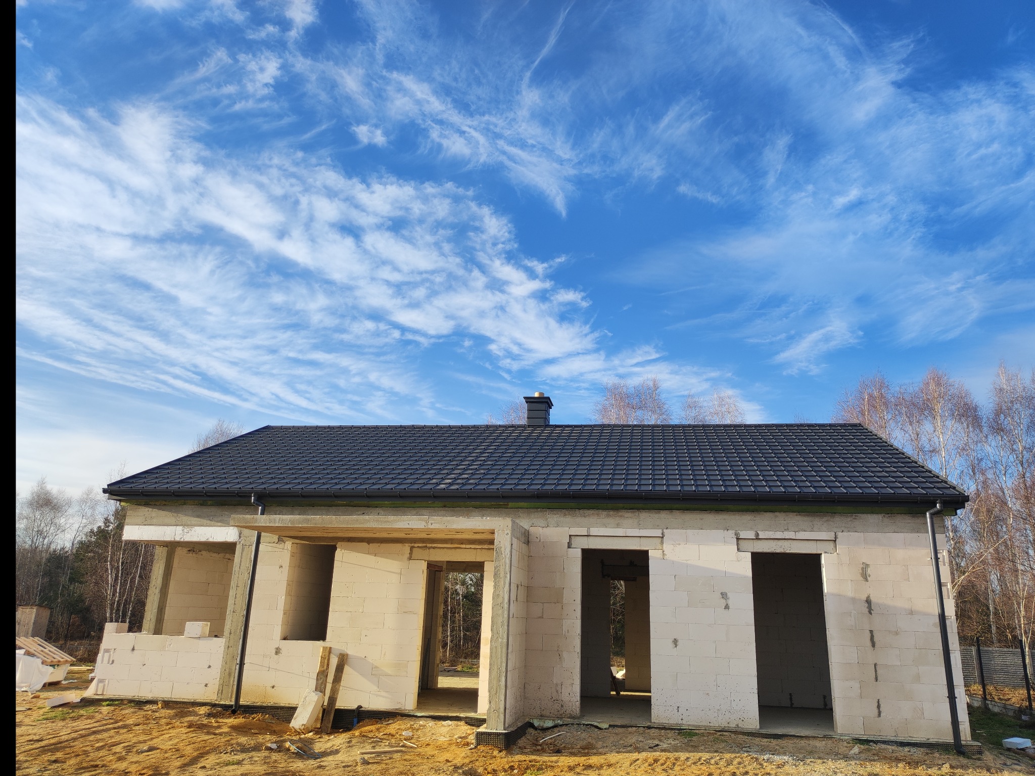 Dom w trakcie budowy z nowym, ciemnym dachem z dachówki ceramicznej, widoczne rynny, ściany z białych pustaków, błękitne niebo z chmurami w tle.