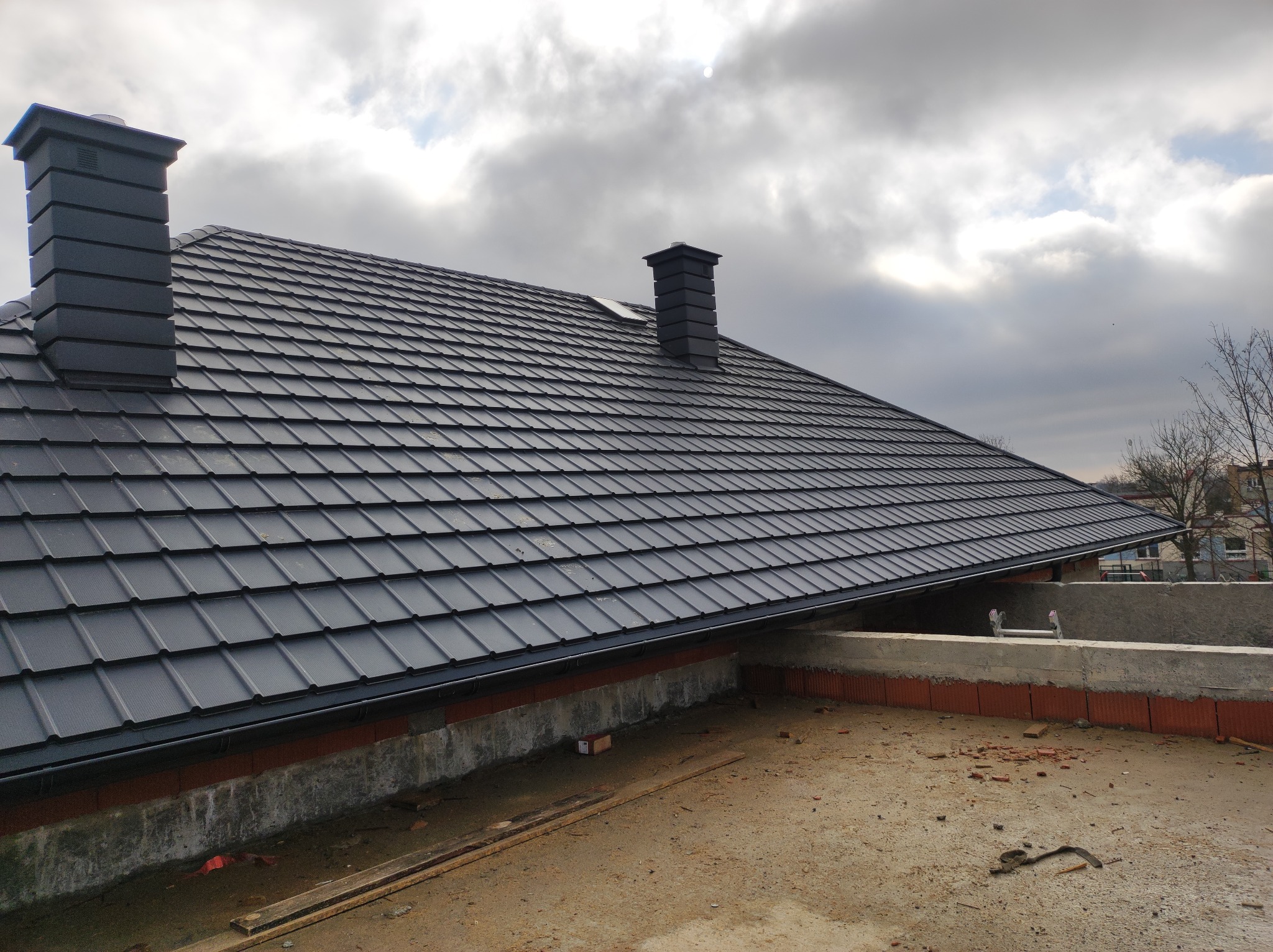Nowy, grafitowy dach z blachodachówki z dwoma kominami w kolorze dachu, widok z poziomu surowego stropu, pochmurne niebo w tle.
