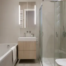 Nowoczesna łazienka w neutralnych barwach z kabiną prysznicową, umywalką z drewnianą szafką, podświetlanym lustrem i wanną z ręcznikiem.