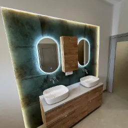 Nowoczesna łazienka z dwoma owalnymi umywalkami nablatowymi, szafką z drewnopodobnym fornirem, podświetlanymi lustrami i zielono-szarą ścianą z efektem patyny.