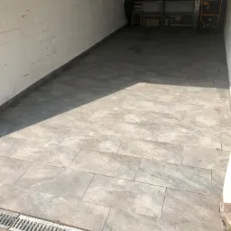 Wyłożona szarymi płytkami podłoga w garażu z odpływem liniowym przy bramie wjazdowej, widoczne metalowe regały z narzędziami w tle.