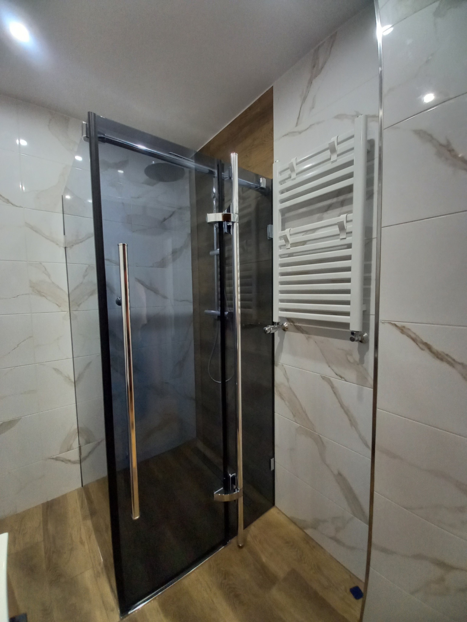 Nowoczesna kabina prysznicowa z grafitowymi profilami i deszczownicą, biały grzejnik drabinkowy na ścianie wyłożonej płytkami imitującymi marmur, drewnopodobna podłoga.