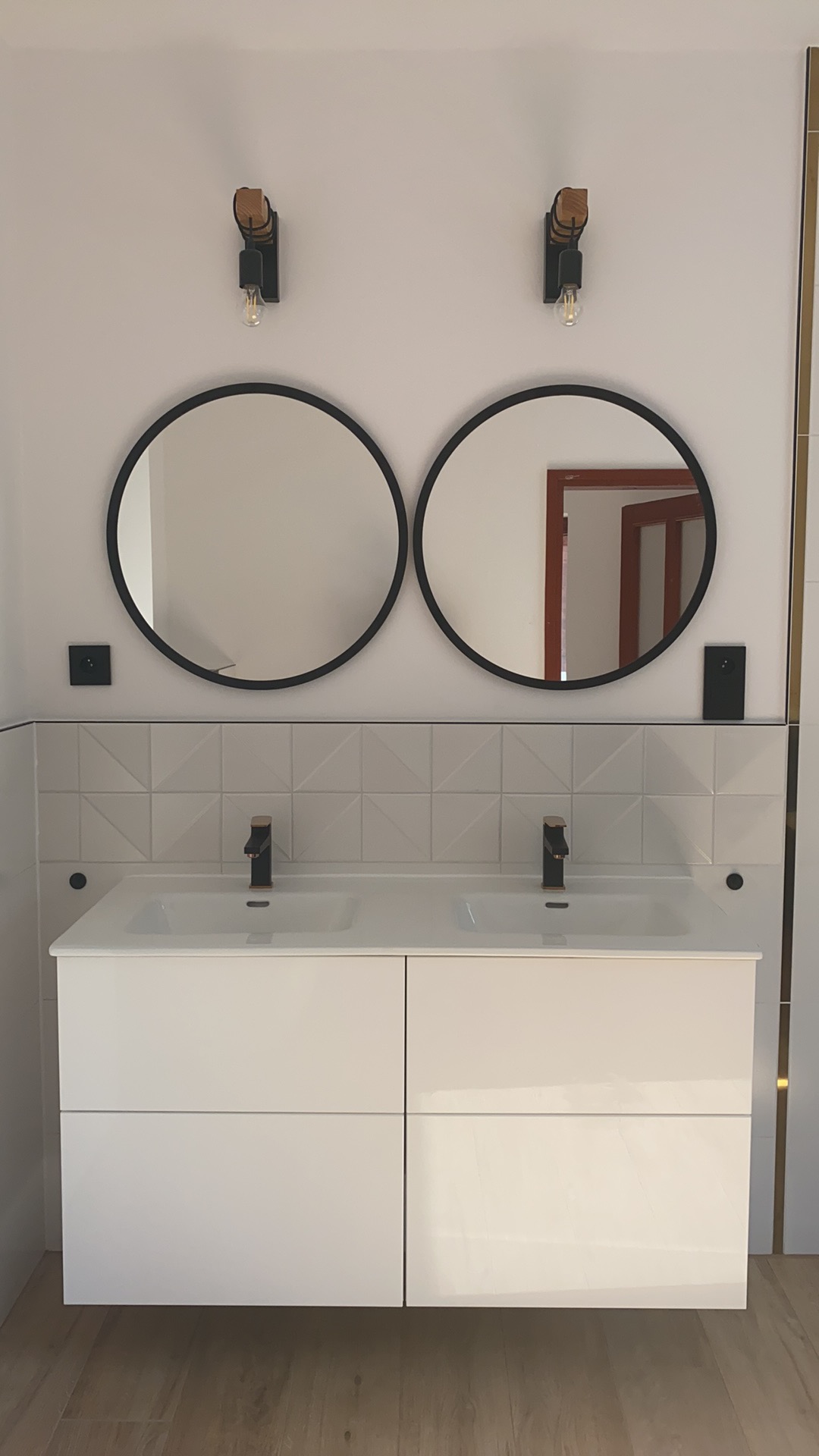 Nowoczesna łazienka z białą szafką pod umywalkę z dwoma czarnymi kranami, dwa okrągłe lustra z czarną ramą i lampami w stylu industrialnym, białe płytki z delikatnym wzorem geometrycznym na ścianie.