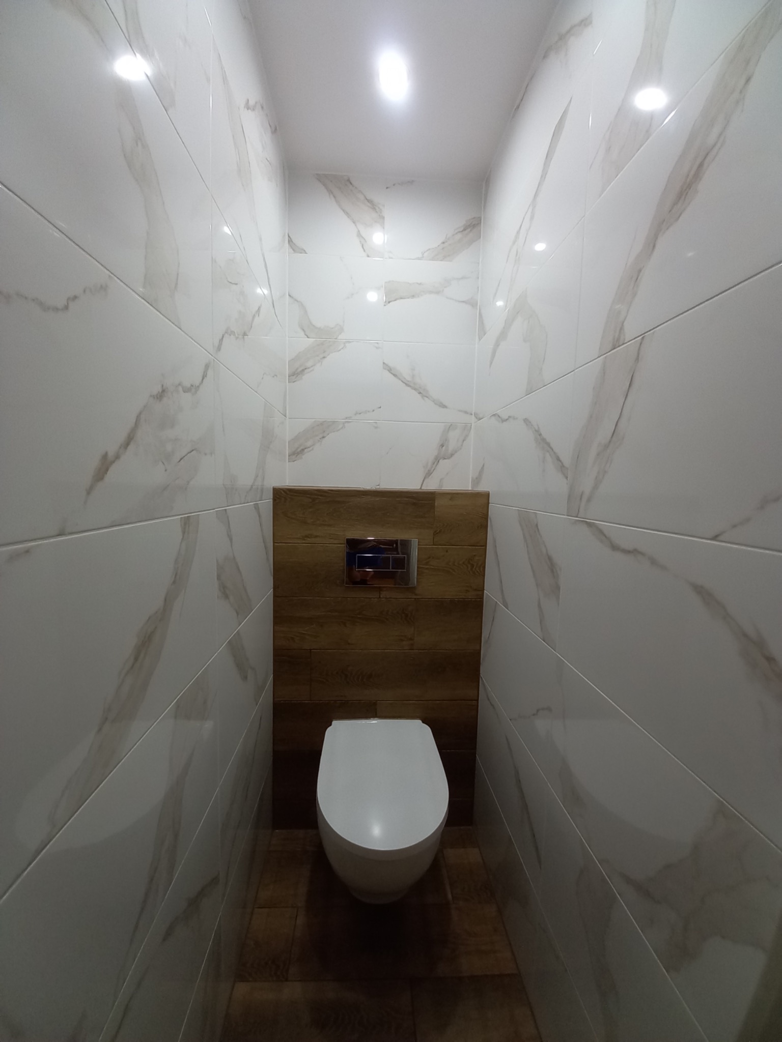 Wąska toaleta z podwieszaną muszlą klozetową, białe płytki imitujące marmur na ścianach, drewnopodobna obudowa stelaża i podłoga z płytek w odcieniach brązu.