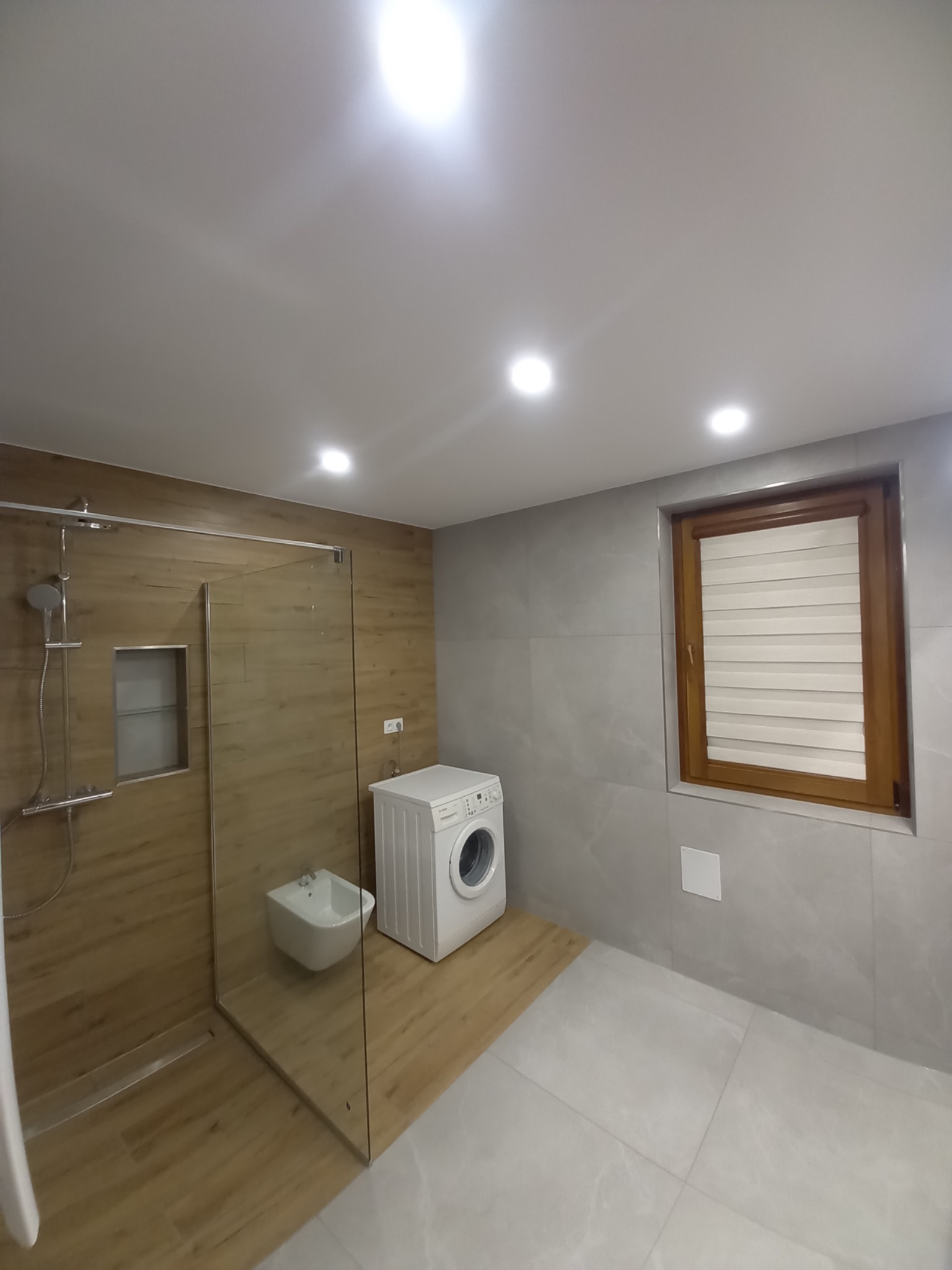 Nowoczesna łazienka po remoncie: kabina prysznicowa typu walk-in z odpływem liniowym i wnęką, bidet, pralka, szare płytki imitujące marmur, drewnopodobna podłoga w strefie prysznica, drewniane okno...