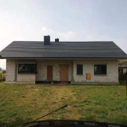 Niedokończony parterowy dom z szarej cegły z ciemną dachówką, oknami z czarnymi ramami i zabitymi deskami drzwiami, na trawiastym terenie.
