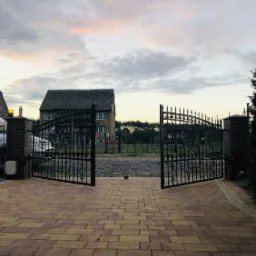 Otwarta, kuta brama wjazdowa z automatyką, prowadząca na posesję z kostką brukową, w tle dom jednorodzinny i zachmurzone niebo o zmierzchu.