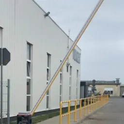 Podniesiona szlabanowa zapora automatyczna przed budynkiem przemysłowym z żółtymi barierkami ochronnymi i stożkiem ostrzegawczym w tle.