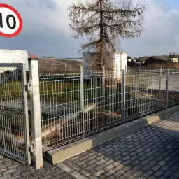 Otwarta brama panelowa z ocynkowanej siatki na betonowej podmur&oacute;wce, widoczny znak ograniczenia prędkości do 10 km/h, teren przemysłowy w tle.