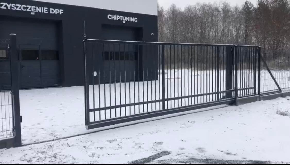 Przesuwna brama wjazdowa z metalowymi prętami na posesji pokrytej śniegiem, na tle budynku z napisem 'CZYSZCZENIE DPF' i 'CHIPTUNING'.