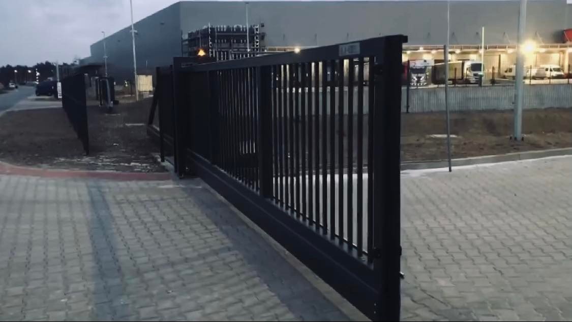 Częściowo otwarta, przesuwna, metalowa brama wjazdowa w kolorze grafitowym, z pionowymi szczeblami, na tle przemysłowego budynku magazynowego i placu manewrowego z zaparkowanymi samochodami...