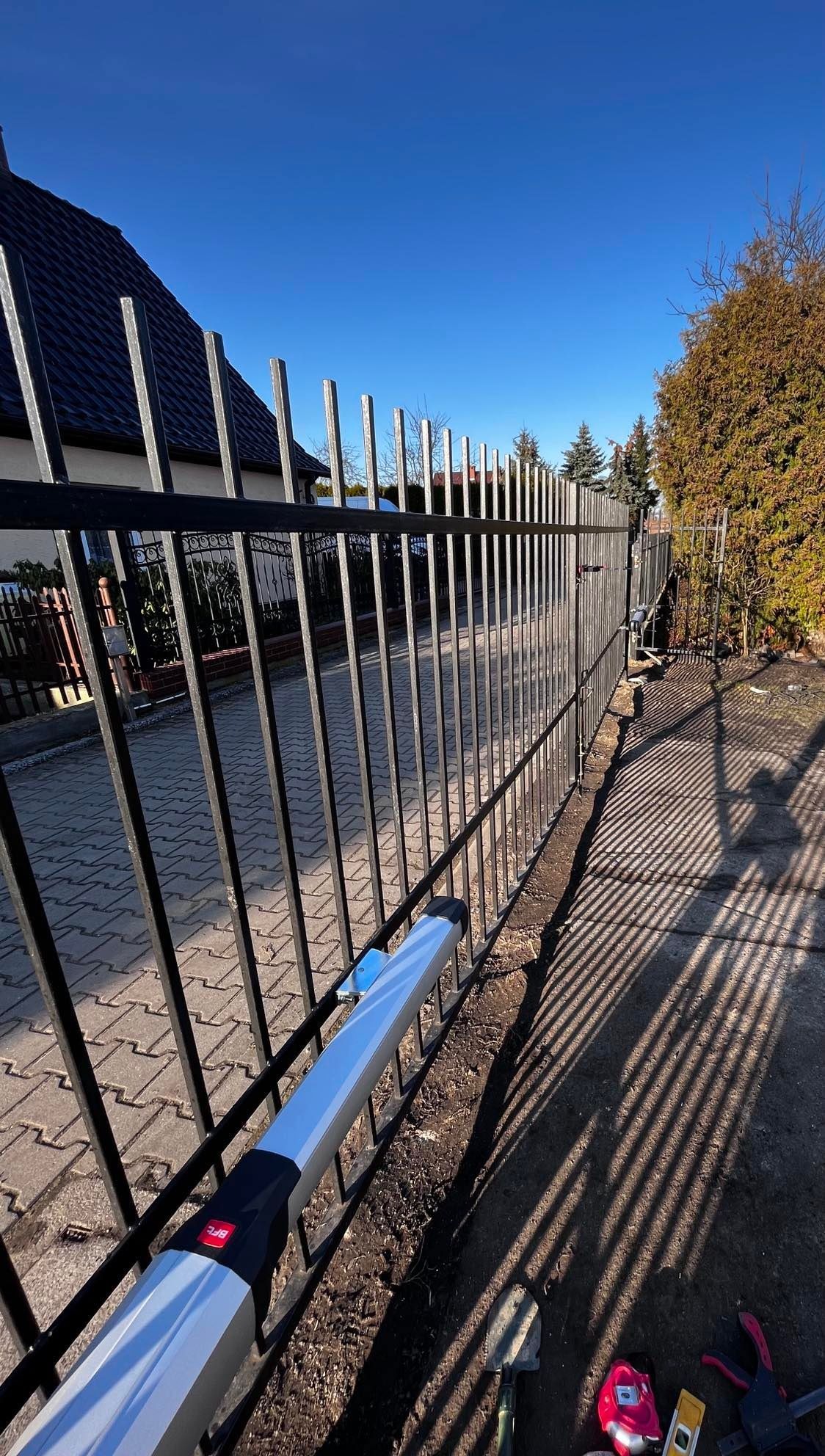 Czarny metalowy płot z automatycznym napędem, widok z niskiej perspektywy, narzędzia montażowe na ziemi, cień rzucany przez ogrodzenie na chodnik.