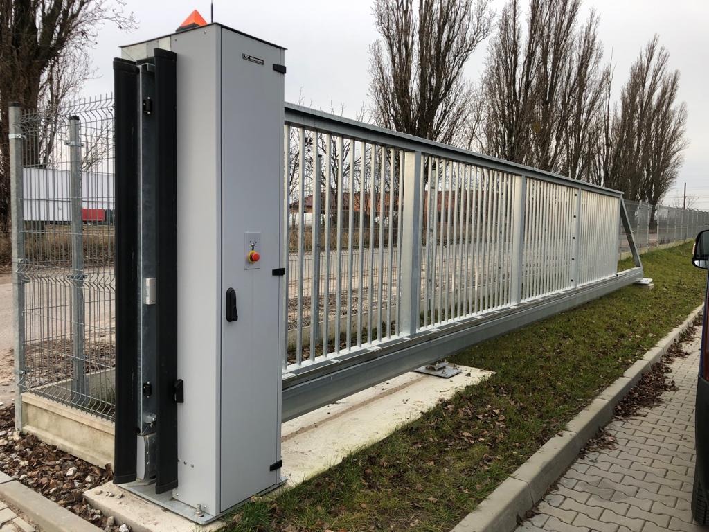 Suwana brama wjazdowa ze stali ocynkowanej, z automatyką, widok z zewnątrz, szara obudowa napędu z przyciskiem awaryjnym, na tle drzew bez liści i ogrodzenia panelowego.