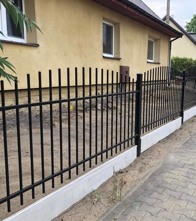 Czarne, pionowe ogrodzenie metalowe na betonowej podmurówce, przylegające do żółtej elewacji budynku w Pile, widoczne z perspektywy chodnika wyłożonego kostką.