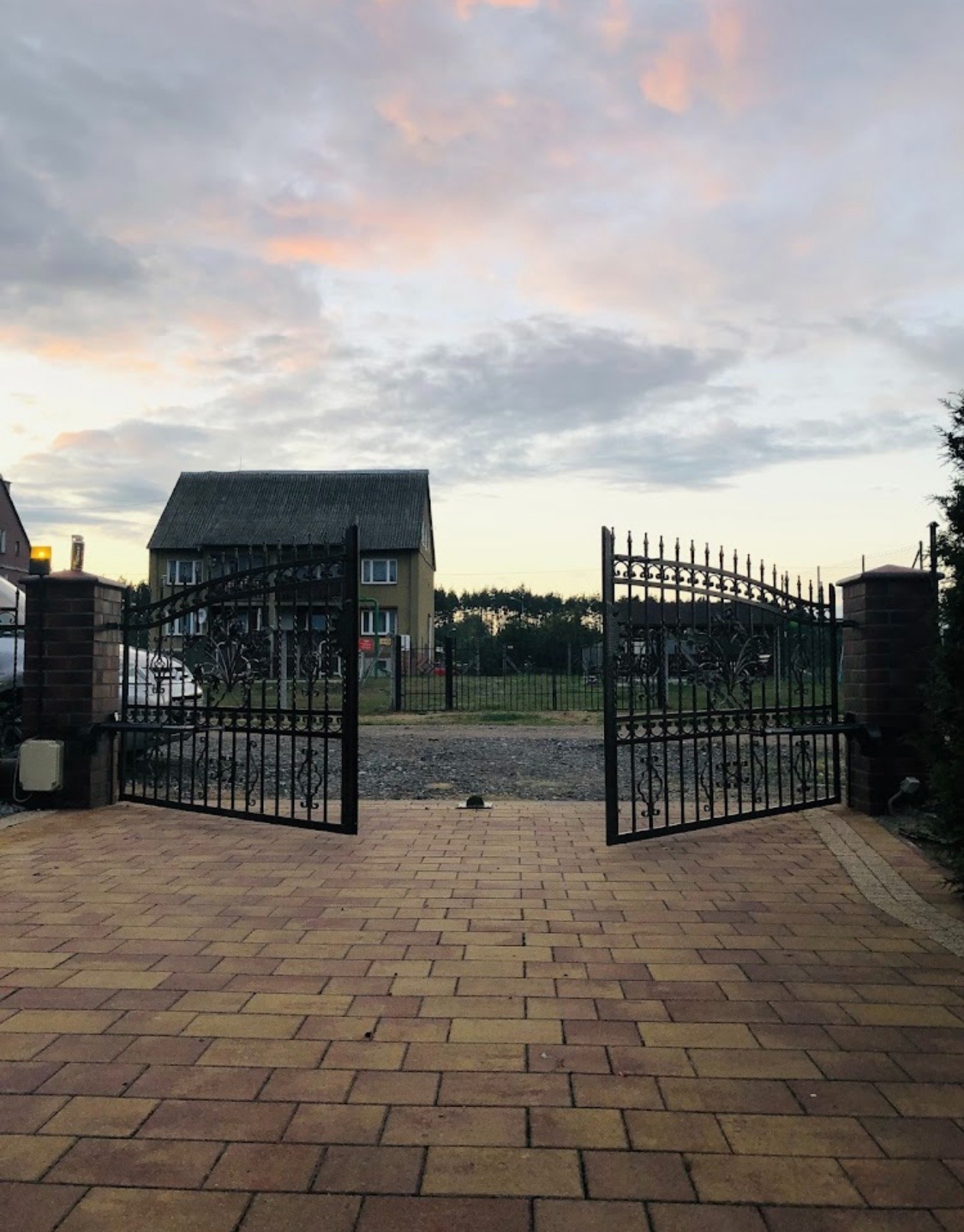 Otwarta, kuta brama wjazdowa z automatyką, prowadząca na posesję z kostką brukową, w tle dom jednorodzinny i zachmurzone niebo o zmierzchu.