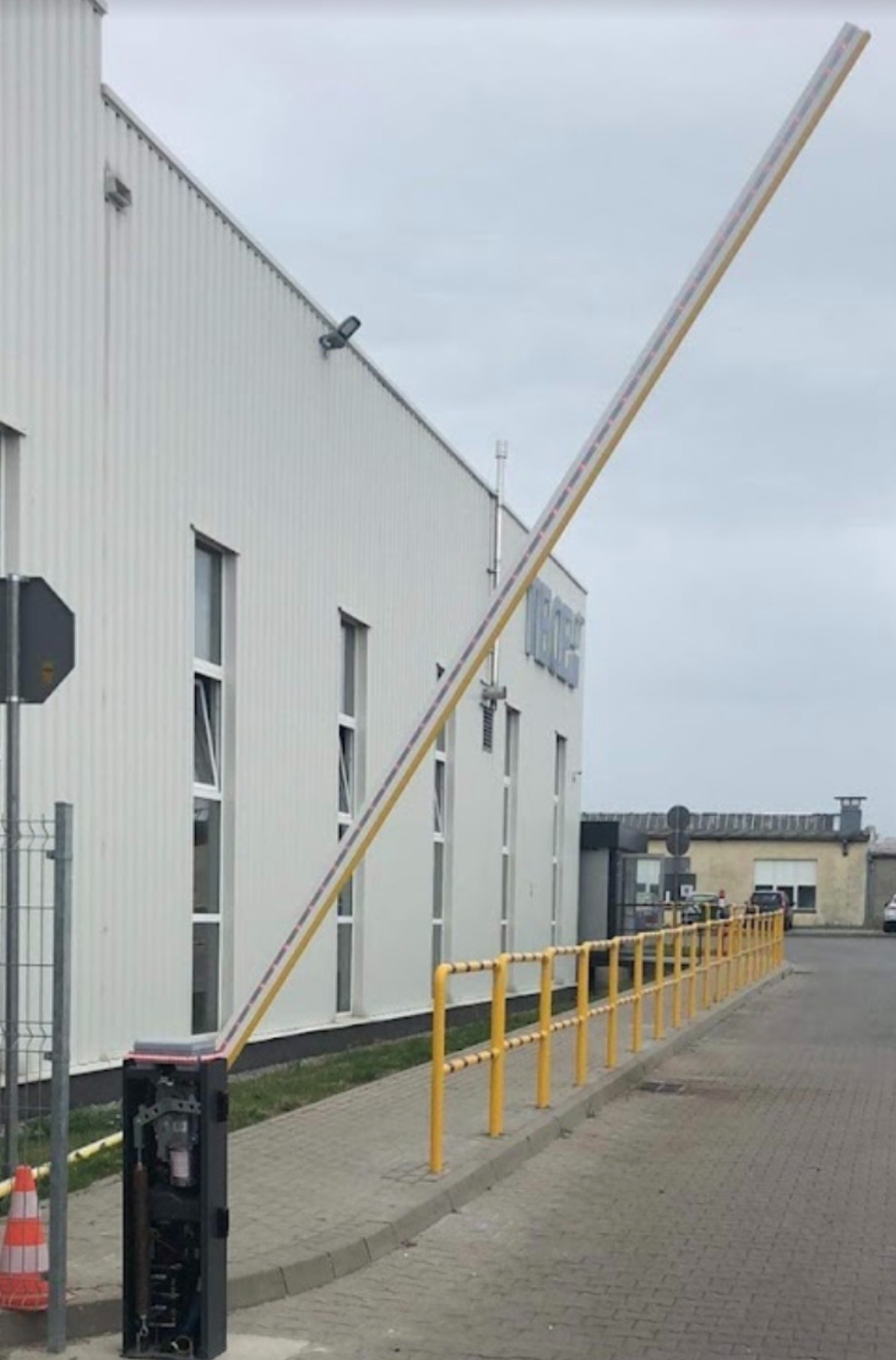 Podniesiona szlabanowa zapora automatyczna przed budynkiem przemysłowym z żółtymi barierkami ochronnymi i stożkiem ostrzegawczym w tle.