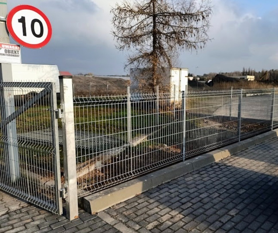 Otwarta brama panelowa z ocynkowanej siatki na betonowej podmurówce, widoczny znak ograniczenia prędkości do 10 km/h, teren przemysłowy w tle.