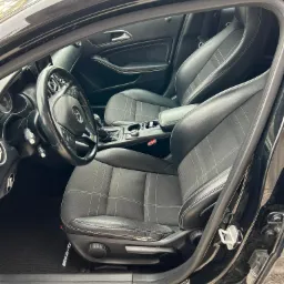 Wnętrze czarnego samochodu marki Mercedes-Benz z widocznymi skórzanymi fotelami z jasną kratką, kierownicą i deską rozdzielczą. Widoczny próg z napisem Mercedes-Benz oraz dywanik z logo AMG.