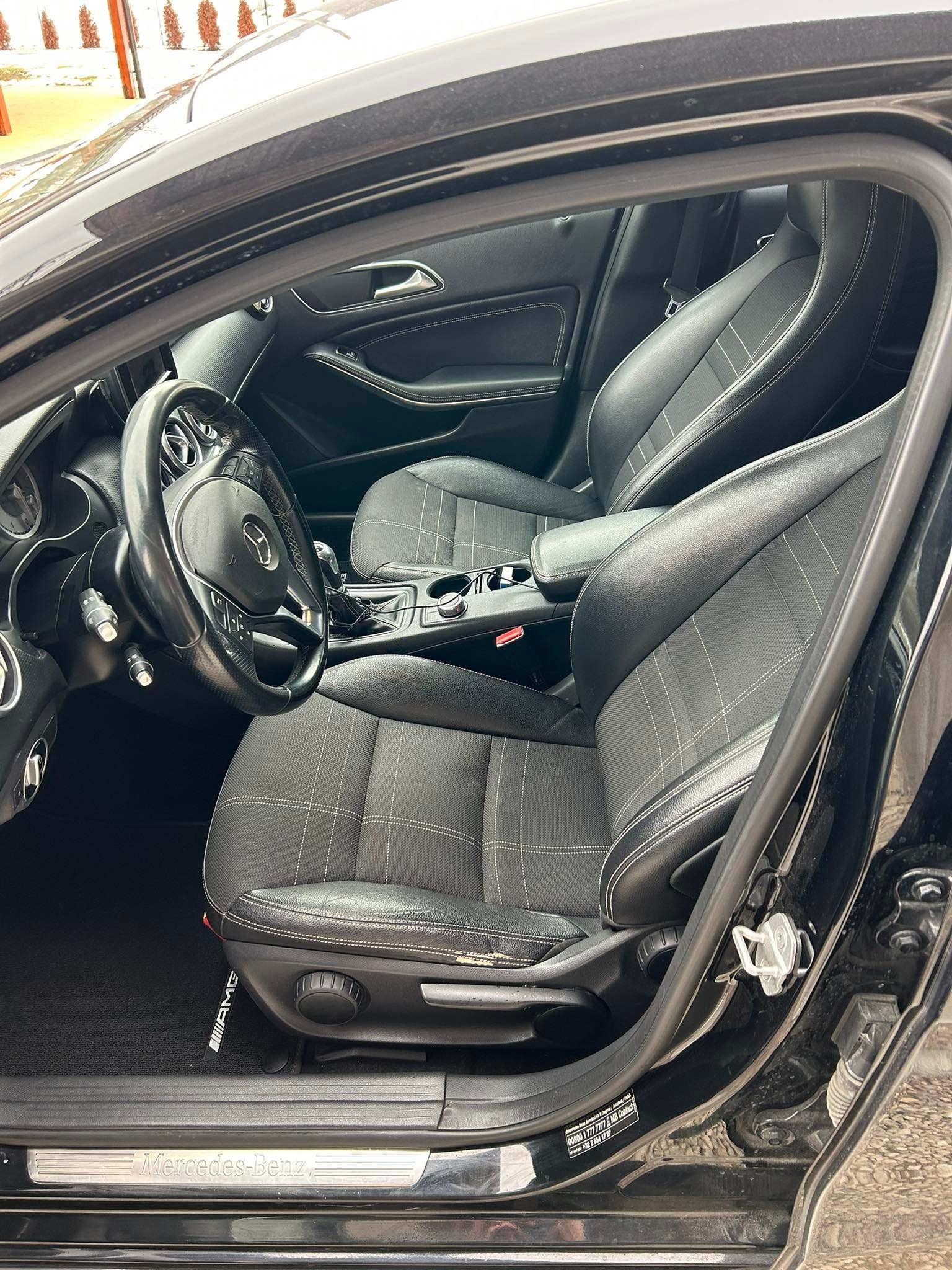 Wnętrze czarnego samochodu marki Mercedes-Benz z widocznymi skórzanymi fotelami z jasną kratką, kierownicą i deską rozdzielczą. Widoczny próg z napisem Mercedes-Benz oraz dywanik z logo AMG.