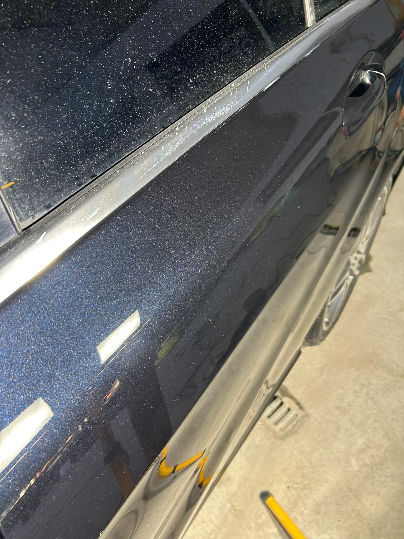Boczny fragment brudnego, ciemnoniebieskiego samochodu z widocznymi zaciekami i zarysowaniami na lakierze, zaparkowanego prawdopodobnie w myjni.