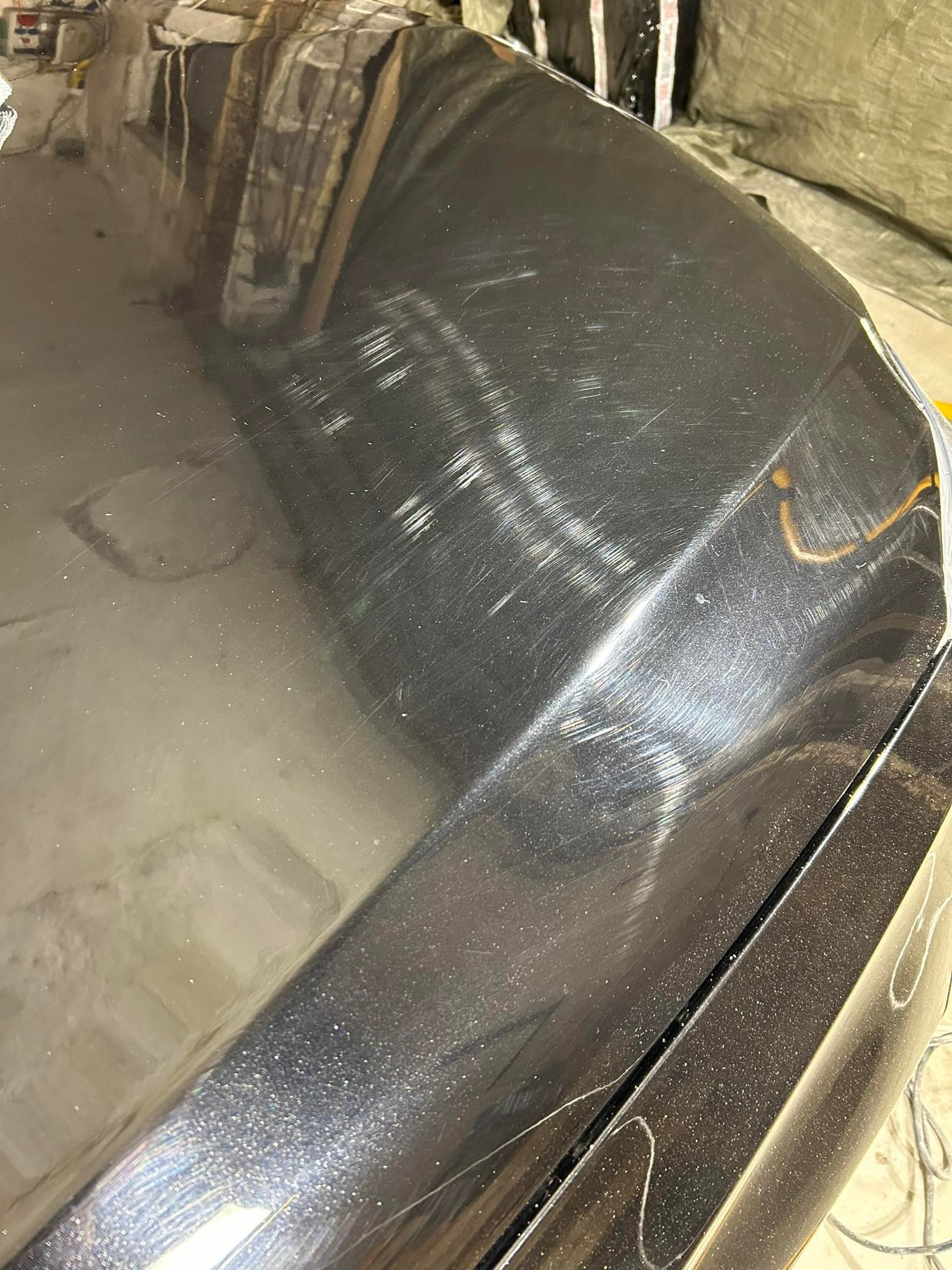 Błyszcząca, czarna maska samochodu z widocznymi zarysowaniami i hologramami po polerowaniu, odblask światła na krawędzi, zabrudzona powierzchnia.