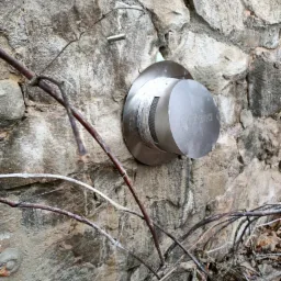 Okucie wentylacyjne ze stali nierdzewnej zamontowane na szarej, kamiennej ścianie zewnętrznej, z widocznymi suchymi gałęziami na pierwszym planie.