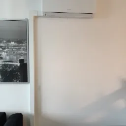 Biała klimatyzacja marki Gree zamontowana na ścianie w mieszkaniu, z widoczną listwą maskującą przewody, obok czarno-białe zdjęcie w ramie.