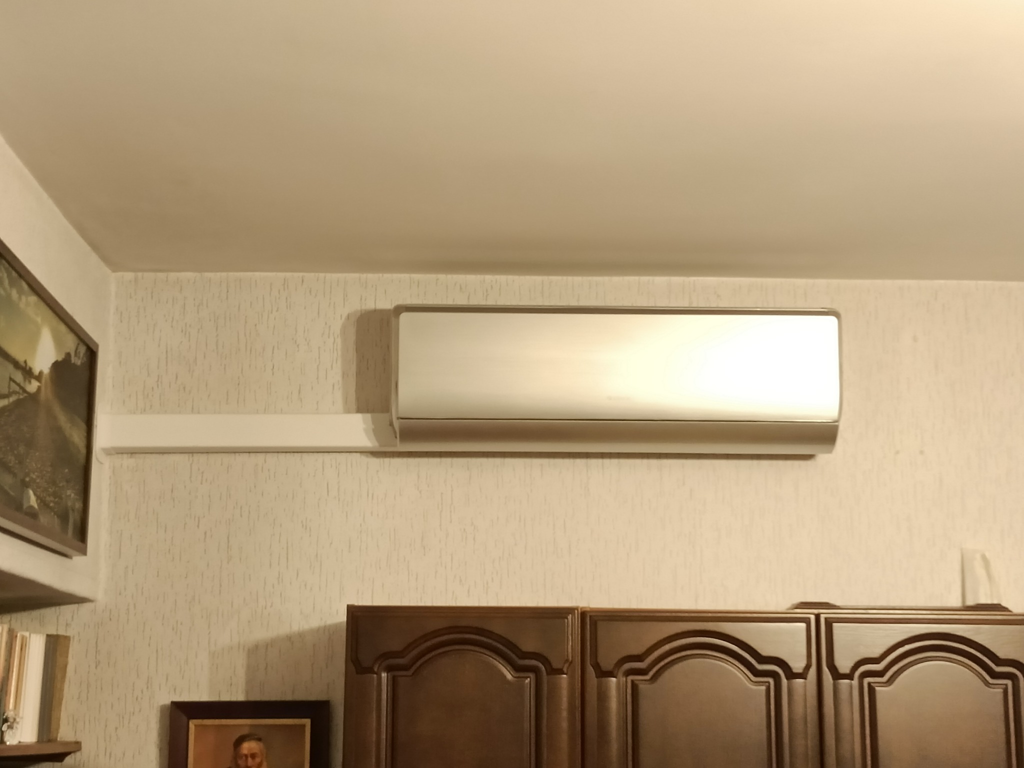 Srebrny klimatyzator ścienny zamontowany na ścianie z tapetą w mieszkaniu, widoczne szafki kuchenne poniżej.