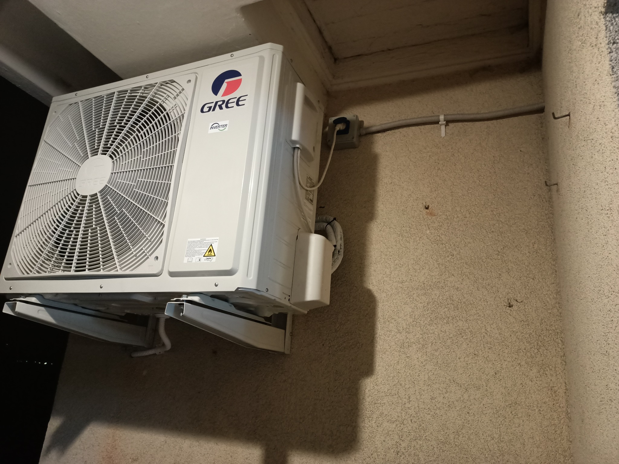 Zewnętrzna jednostka klimatyzacji marki Gree zamontowana na chropowatej, beżowej ścianie budynku, z widocznymi elementami instalacji elektrycznej w szarej osłonie.