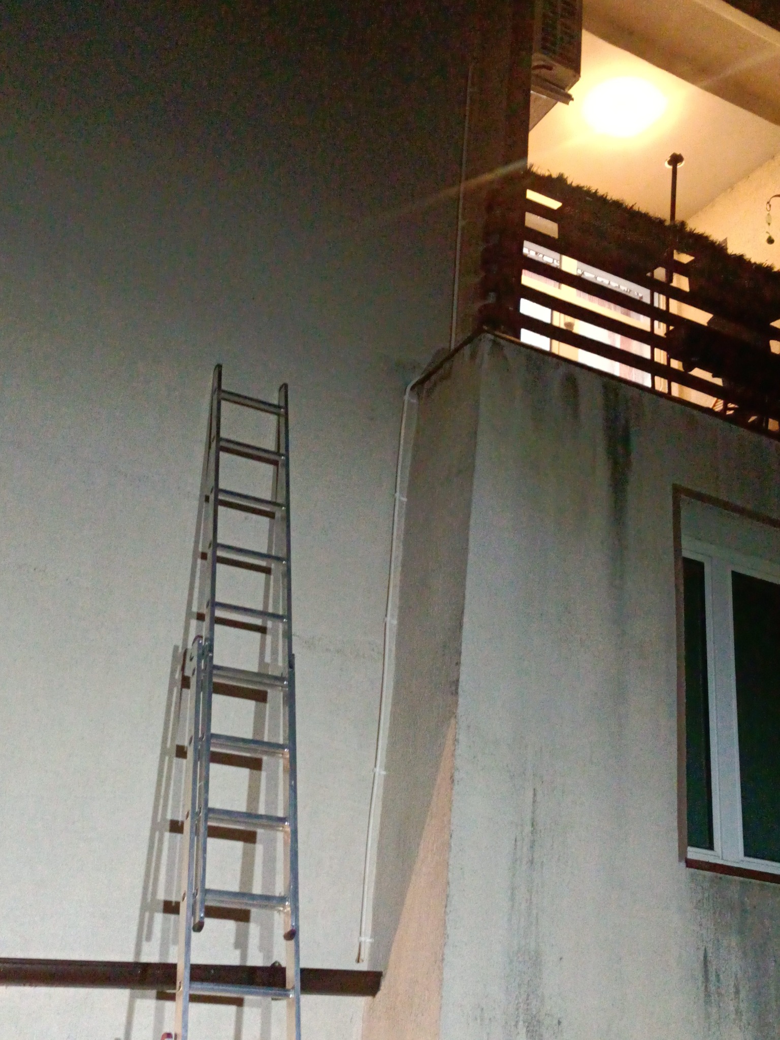 Aluminiowa drabina oparta o ścianę budynku z balkonem, widoczna instalacja klimatyzacji na balkonie, ujęcie nocne.