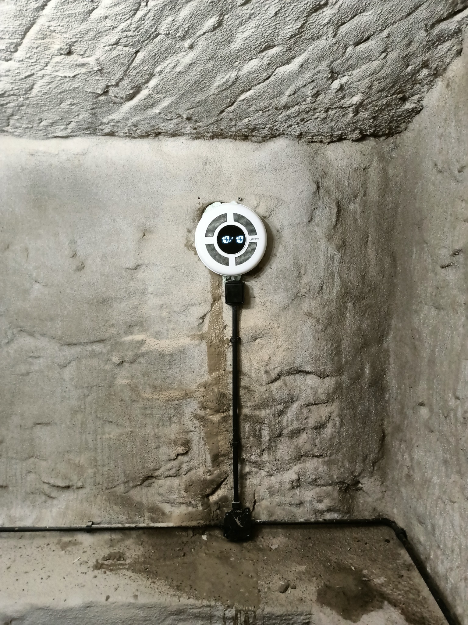 Okrągły, biały czujnik z wyświetlaczem przymocowany do szarej, nierównej ściany w piwnicy. Widoczne okablowanie i instalacja w narożniku pomieszczenia.