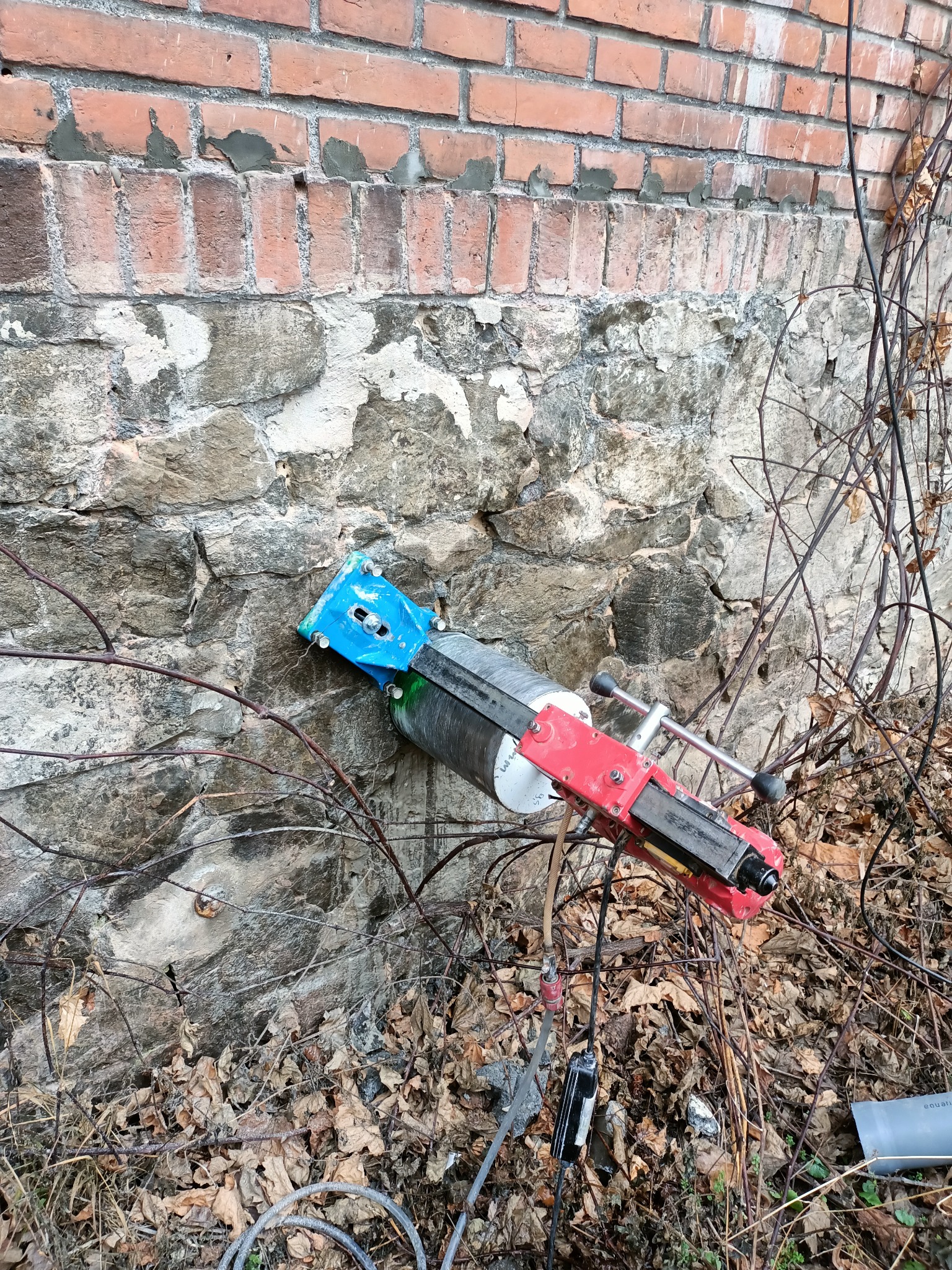 Wiertnica rdzeniowa z czerwoną ramą i niebieskim uchwytem zamocowana do starej, kamienno-ceglanej ściany zewnętrznej budynku, otoczona suchymi gałęziami i opadłymi liśćmi.