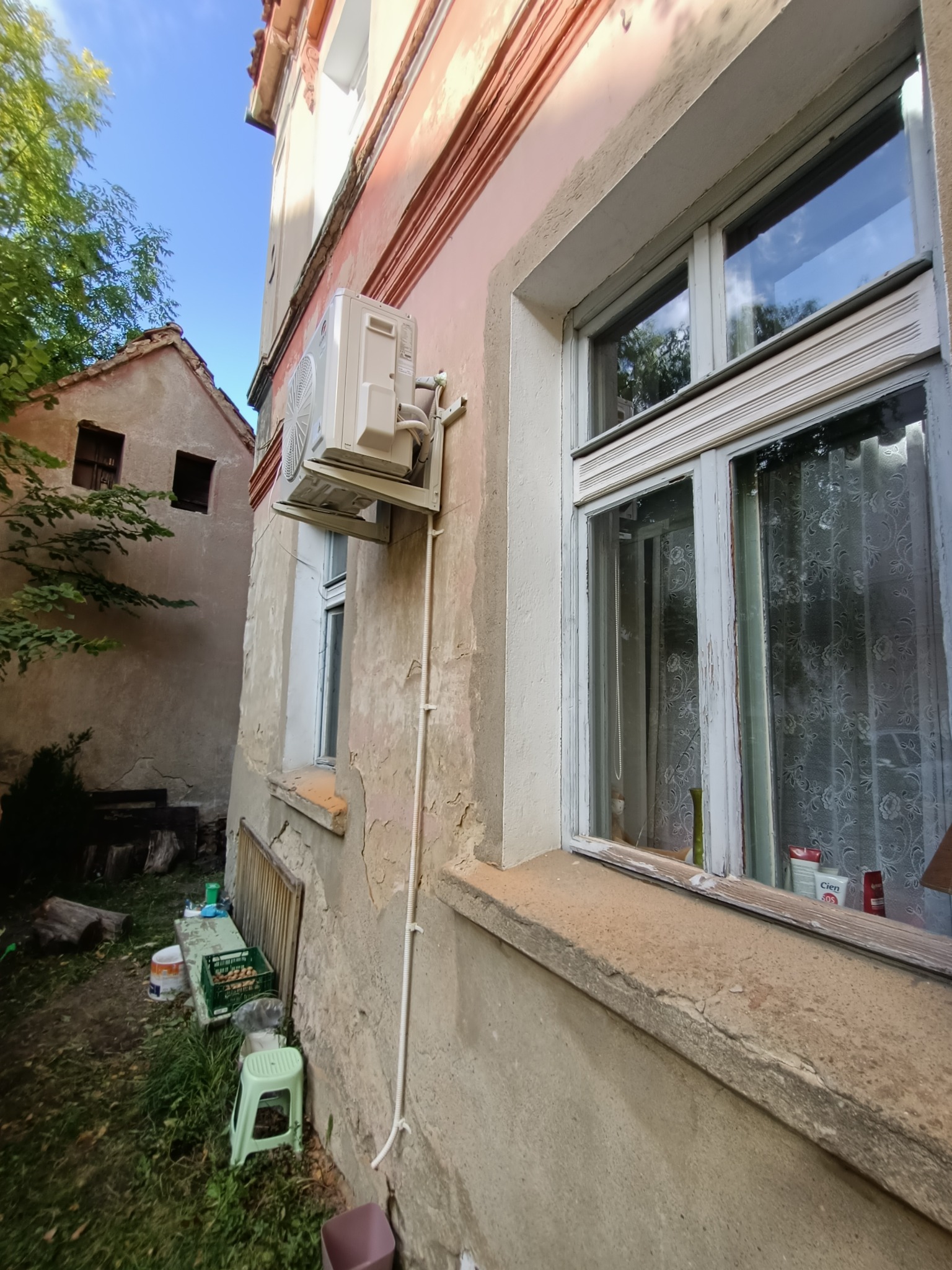 Zewnętrzna jednostka klimatyzacji zamontowana na elewacji starego budynku z oknem z białymi ramami, widok z dołu