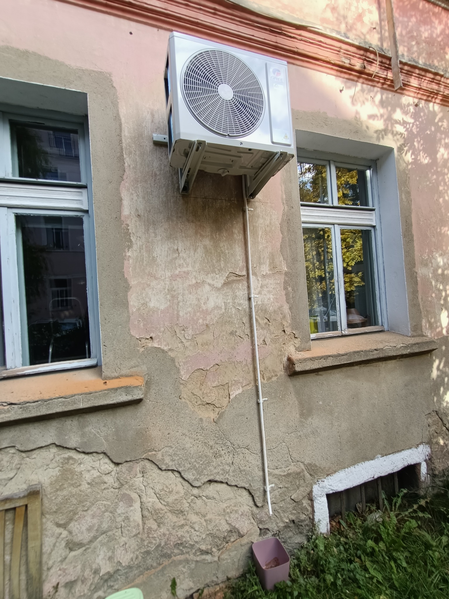 Zewnętrzna jednostka klimatyzacji zamontowana na ścianie starego budynku z odpadającym tynkiem, widoczna rurka odprowadzająca skropliny i okna z odbiciami drzew.