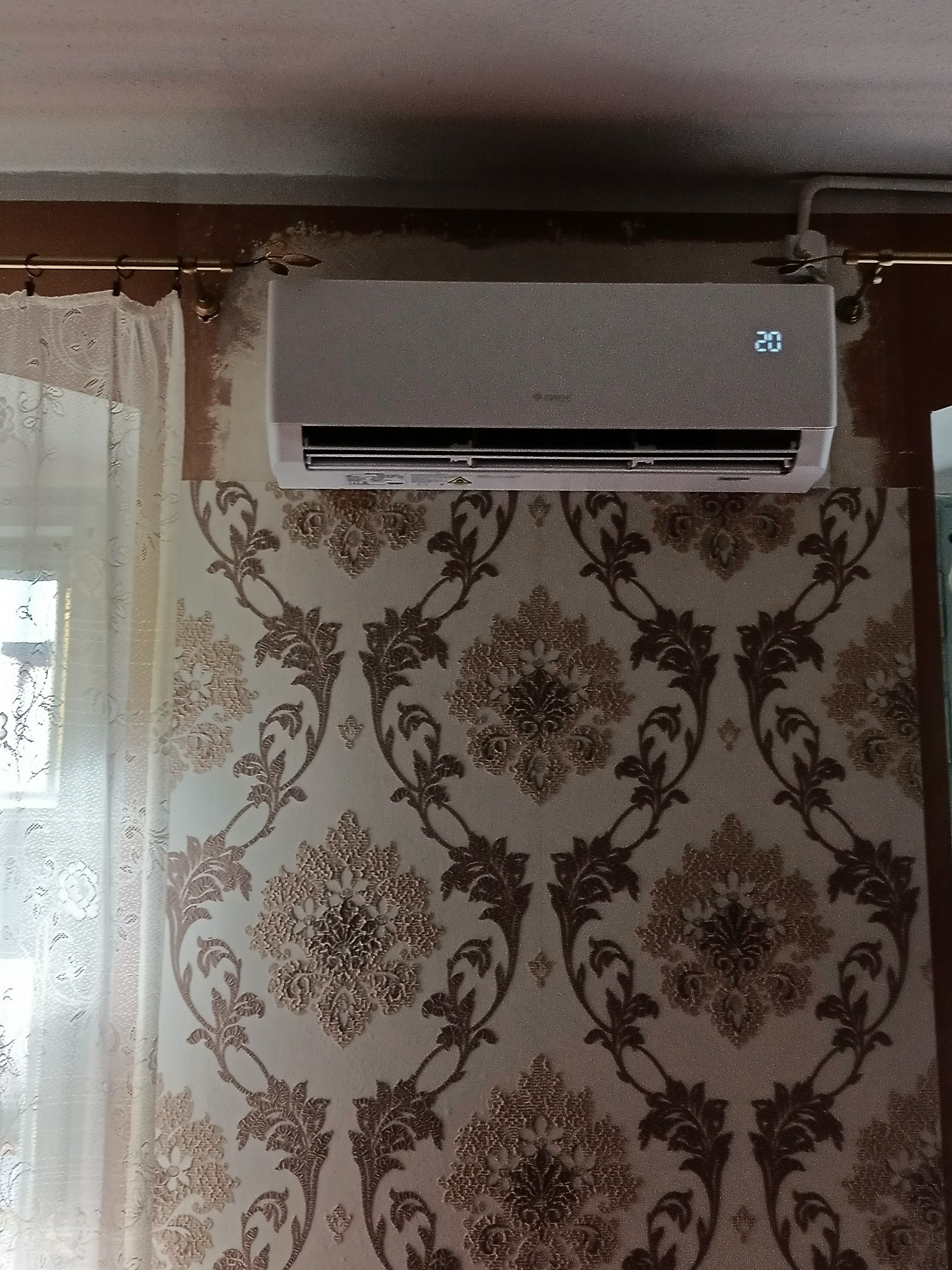 Biała jednostka klimatyzacyjna zamontowana na ścianie z wzorzystą tapetą w odcieniach brązu i beżu, z widocznym oknem z firanką po lewej stronie.