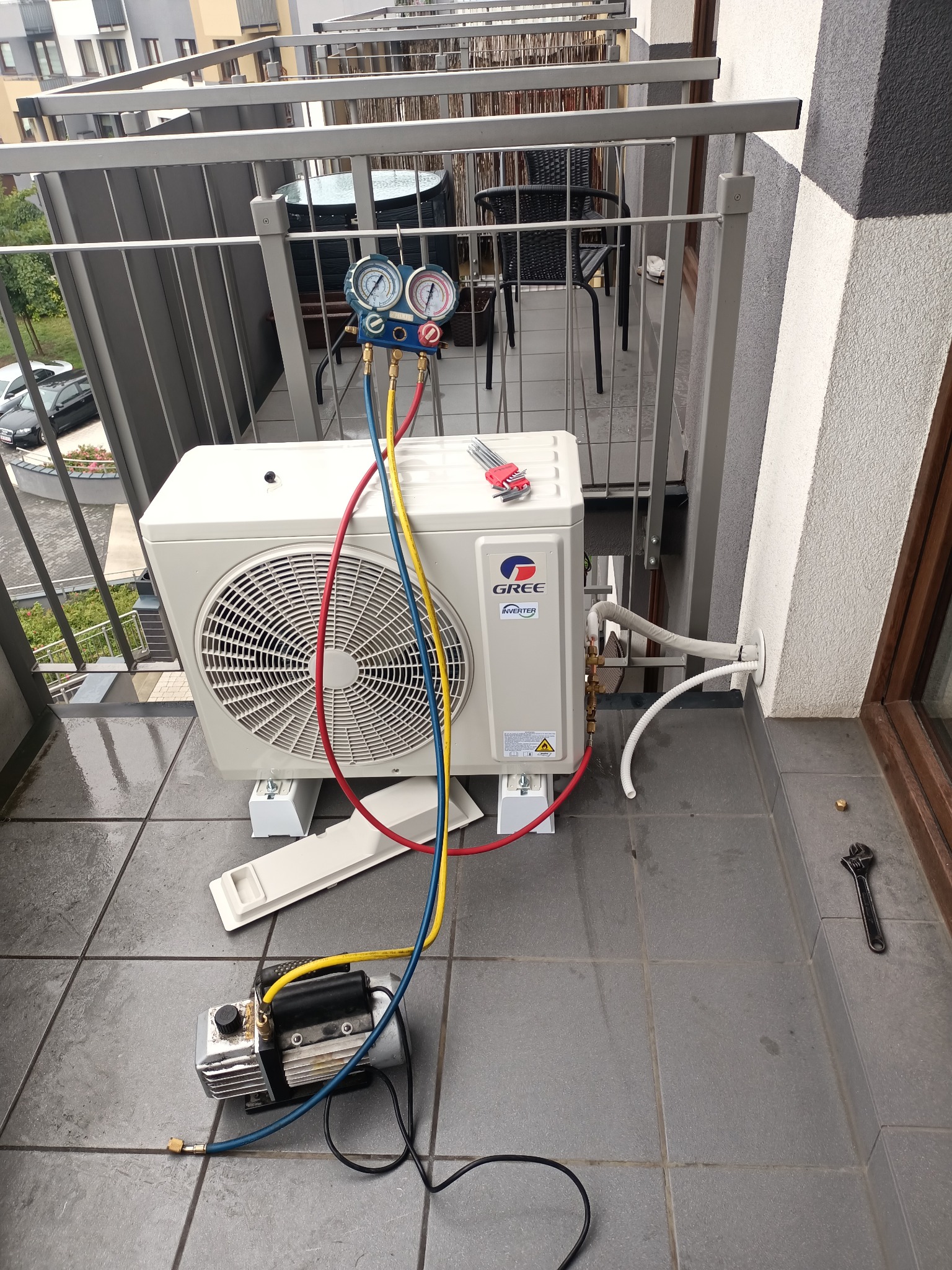 Jednostka zewnętrzna klimatyzacji marki Gree, podłączona do pompy próżniowej i manometrów, umieszczona na balkonie, widoczne narzędzia i kable.