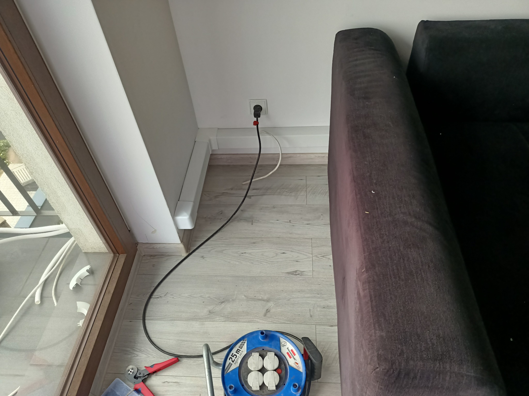 Przedłużacz zwinięty na bębnie leżący na podłodze obok okna i sofy, kabel podłączony do gniazdka elektrycznego w listwie przypodłogowej. Widoczne narzędzia do pracy.