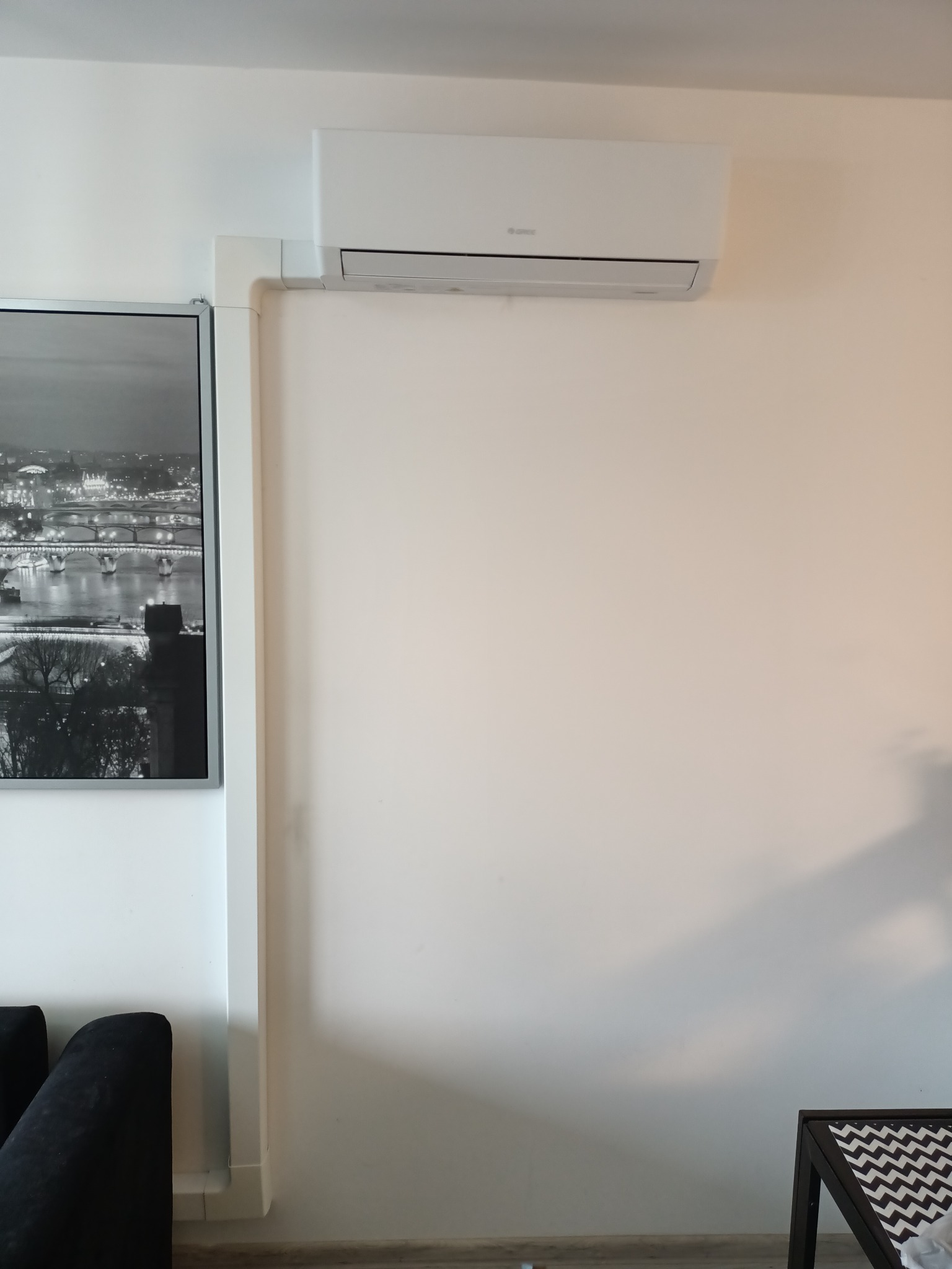 Biała klimatyzacja marki Gree zamontowana na ścianie w mieszkaniu, z widoczną listwą maskującą przewody, obok czarno-białe zdjęcie w ramie.