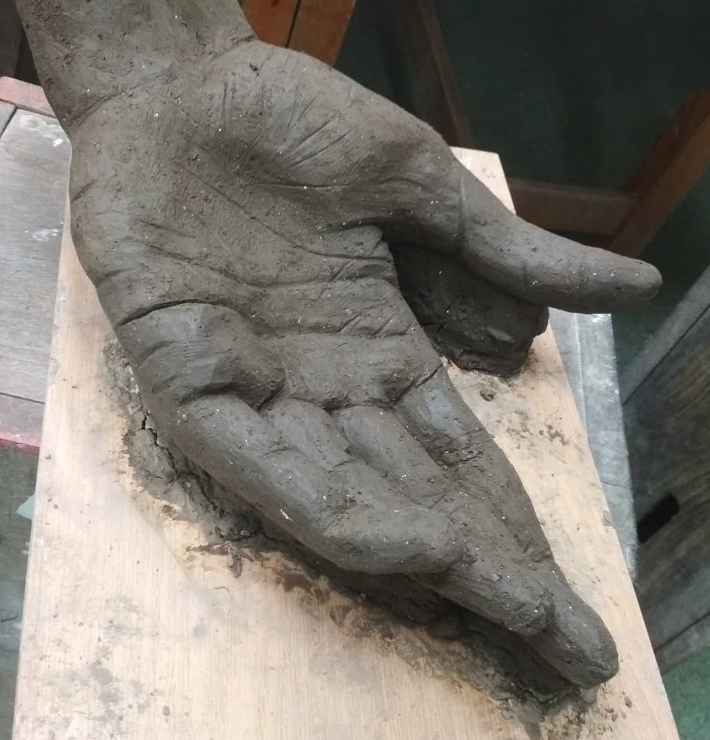 Gliniana rzeźba dłoni w trakcie tworzenia, leżąca na drewnianej desce, widoczne ślady pracy i niedoskonałości detali.