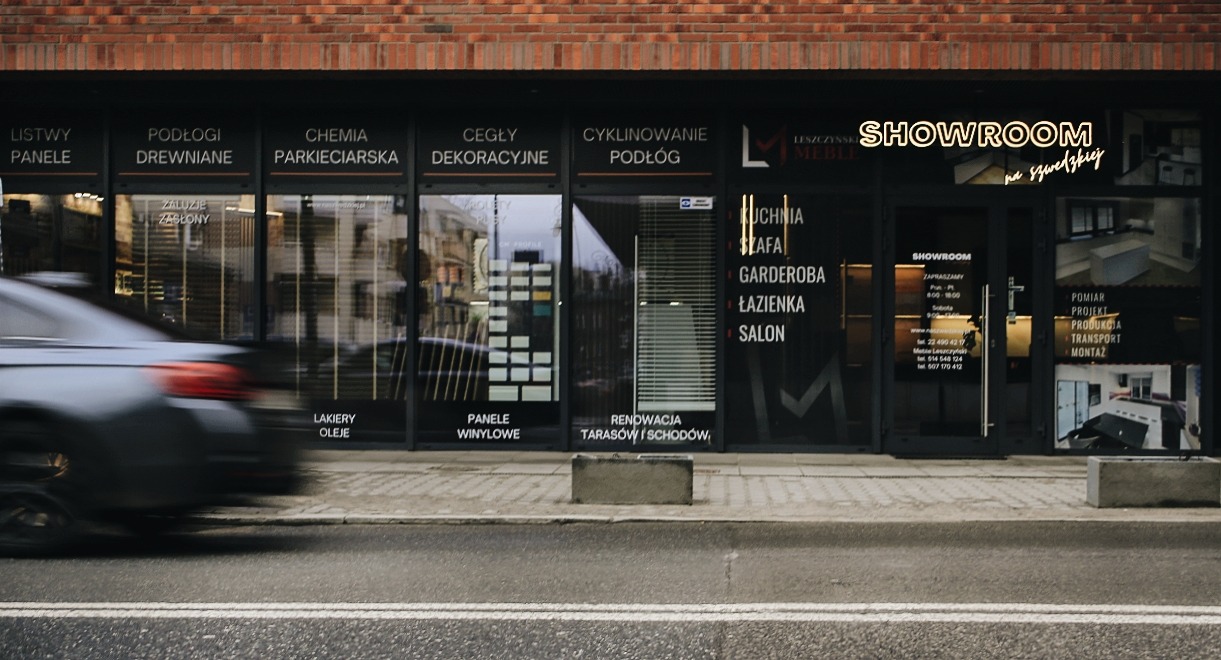 Fasada sklepu z podłogami i meblami, widoczny ruch uliczny, w tle ceglana ściana budynku.