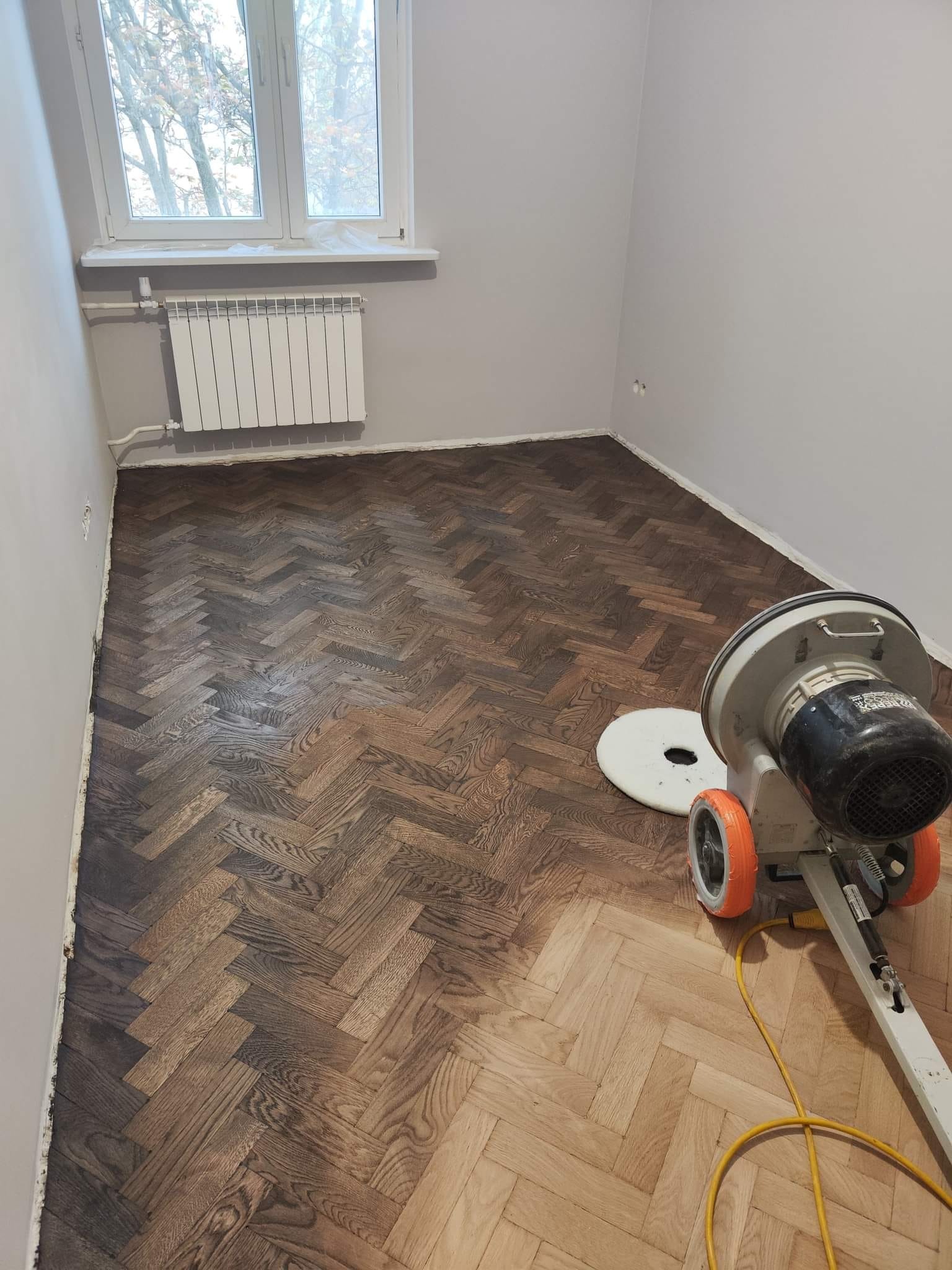 Pokój z parkietem ułożonym w jodełkę, częściowo odnowionym, z widoczną maszyną do cyklinowania z pomarańczowymi kołami i żółtym kablem.