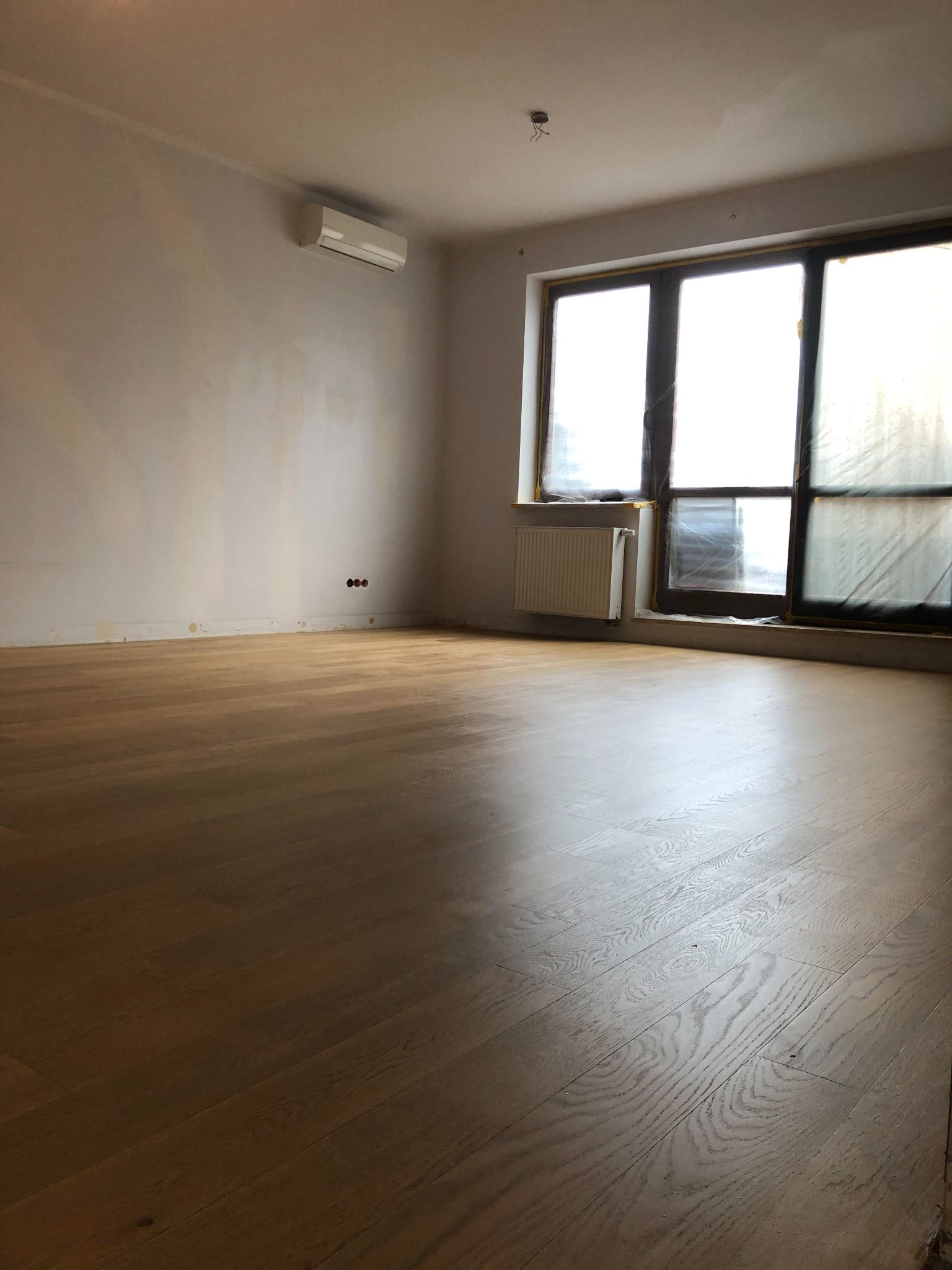 Jasne, drewniane deski podłogowe w pustym pokoju z dużym oknem zabezpieczonym folią, widoczny grzejnik i klimatyzator na ścianie, ściany w trakcie malowania.