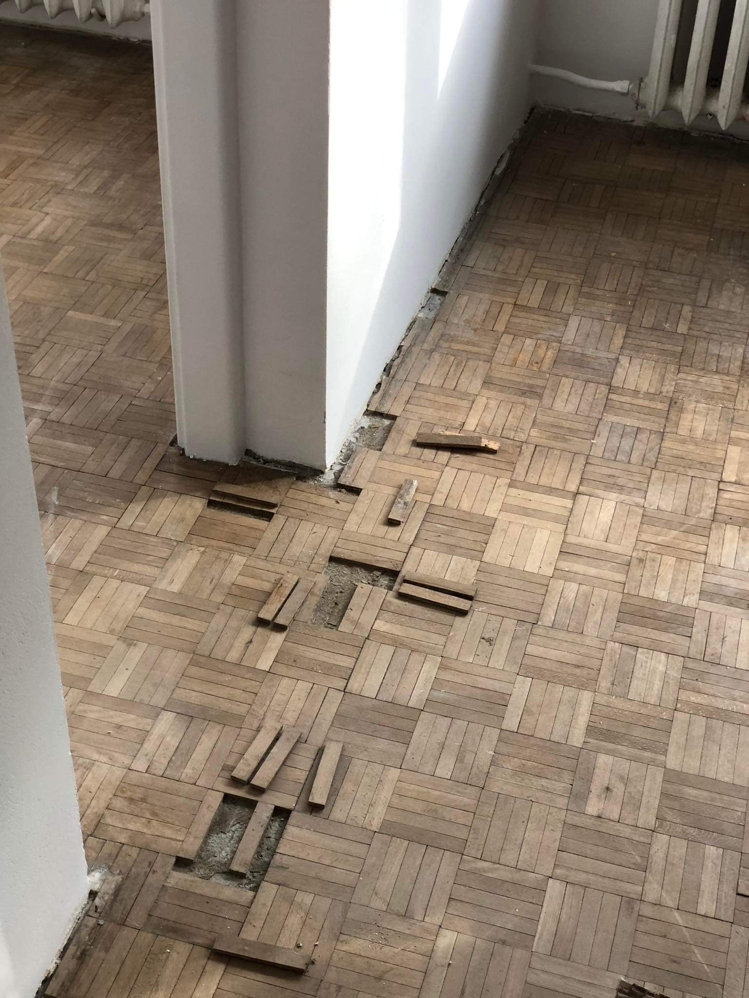 Zniszczona, stara podłoga parkietowa z ubytkami i odspojonymi klepkami, widoczne fragmenty podłoża, biała ściana w tle.