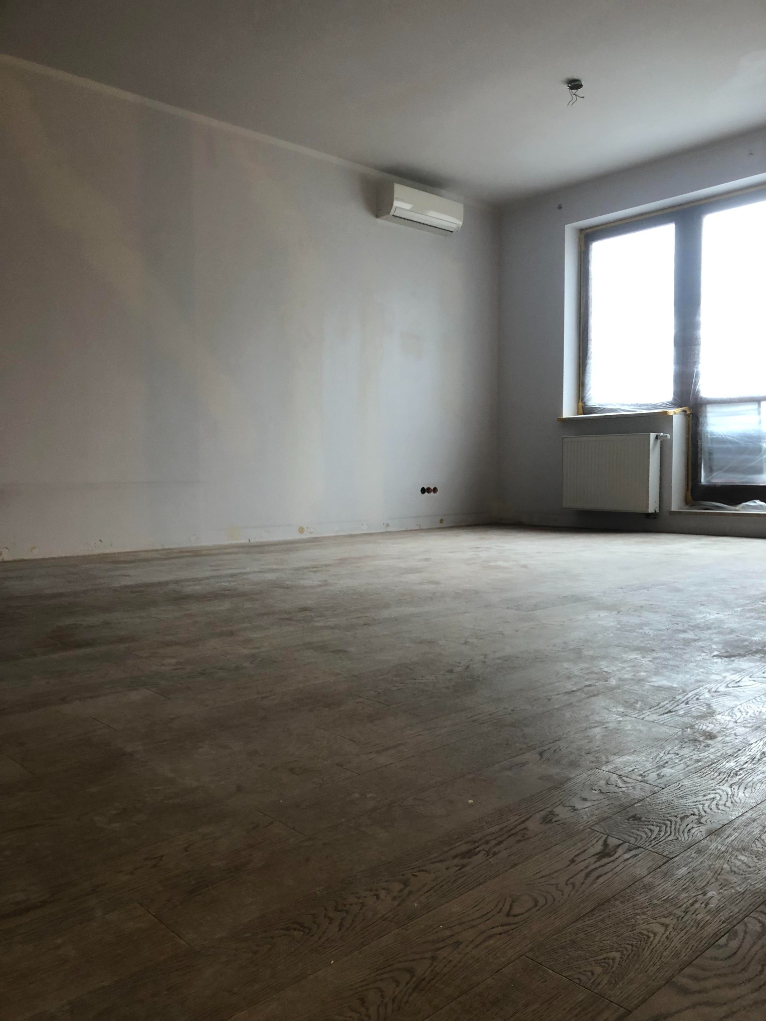 Pokój po renowacji podłogi z widocznymi deskami z wyraźnym usłojeniem, ściany przygotowane do malowania, okno z grzejnikiem, klimatyzator na ścianie, widoczne gniazdka elektryczne.