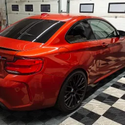 Czerwone BMW M2 z przyciemnionymi szybami, zaparkowane w garażu z podłogą w szachownicę, widok z tyłu i boku, tablica rejestracyjna 'Black Mirror'.