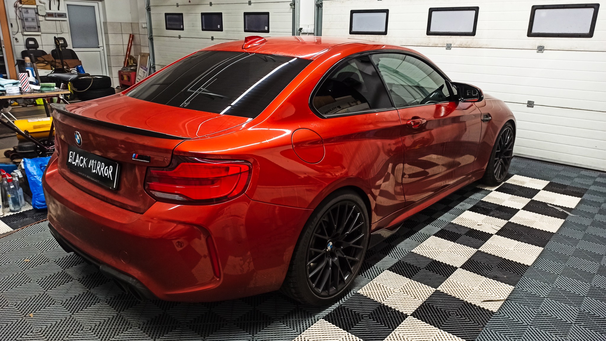 Czerwone BMW M2 z przyciemnionymi szybami, zaparkowane w garażu z podłogą w szachownicę, widok z tyłu i boku, tablica rejestracyjna 'Black Mirror'.
