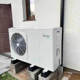 Biała jednostka zewnętrzna z logo Midea, zamontowana na betonowych blokach fundamentowych przy białej ścianie budynku z brązowymi drzwiami i oknem.