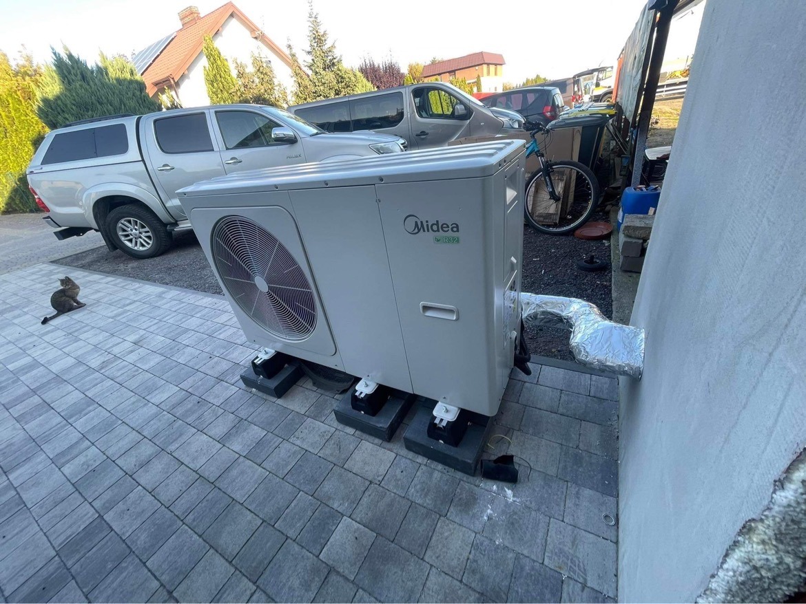 Zewnętrzna jednostka pompy ciepła Midea zamontowana na betonowej kostce brukowej, z widocznym kotem siedzącym obok i fragmentem zabudowań w tle.