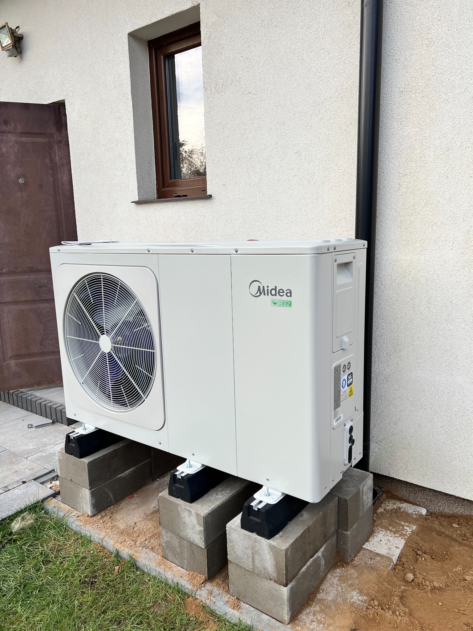Biała jednostka zewnętrzna z logo Midea, zamontowana na betonowych blokach fundamentowych przy białej ścianie budynku z brązowymi drzwiami i oknem.