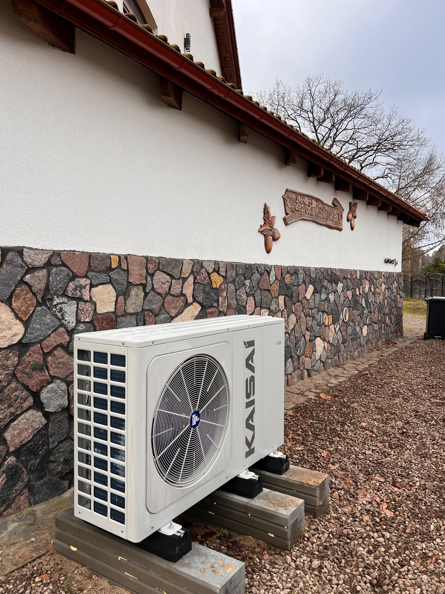 Biała zewnętrzna jednostka pompy ciepła Kaisai, umieszczona na betonowych blokach, przed budynkiem z elewacją z kamienia i białego tynku, w tle drzewo bez liści.
